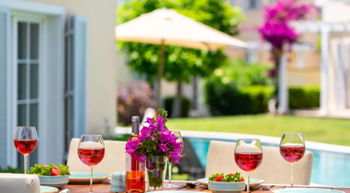 Table set with glasses of rosé wine, salad, and flowers, with a pool and greenery in the background.