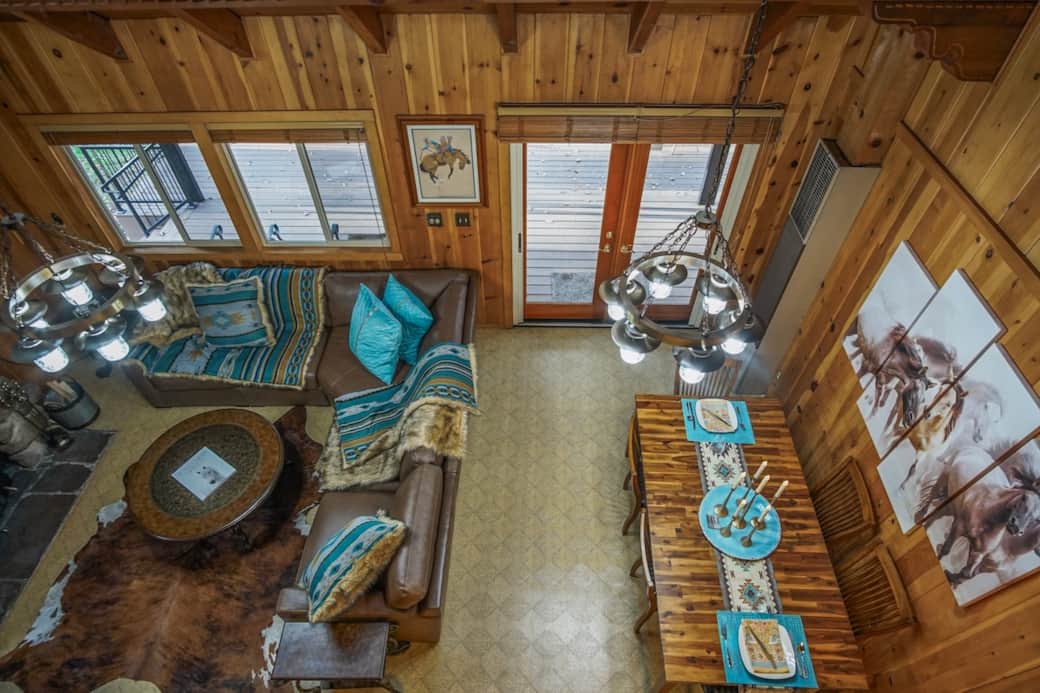 Loft-level view of the great room—featuring soaring wood ceilings, statement chandeliers, and plush leather seating. Bold turquoise textiles tie the open living and dining spaces together, with the outdoor deck just beyond the glass doors.