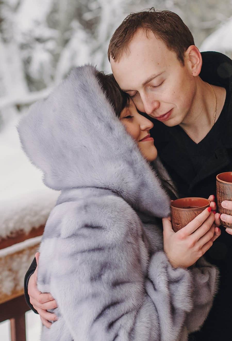 Couple sharing warm drinks on a snowy log cabin deck in winter