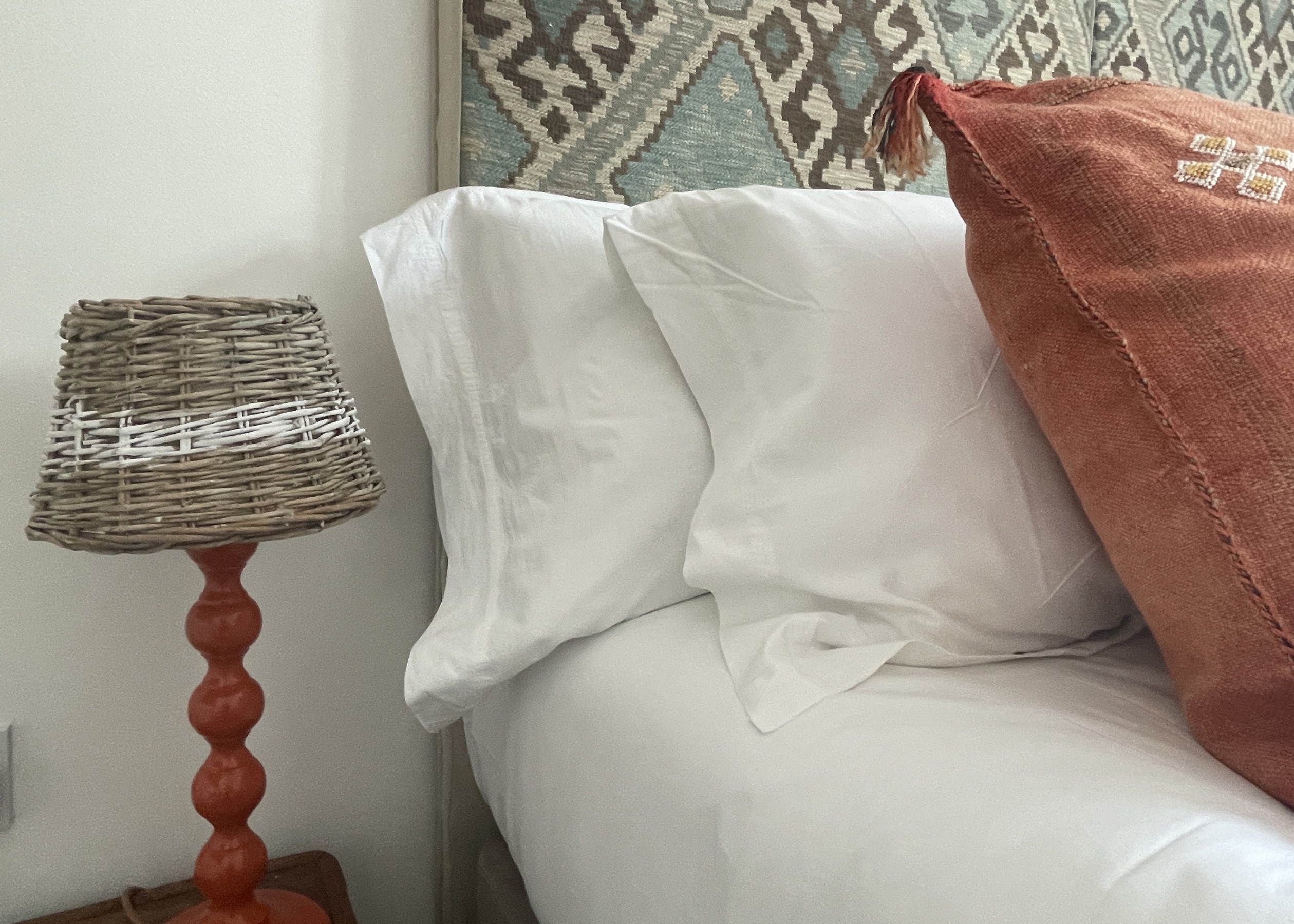 White bedlinen with orange Moroccan cushion and artisan bobbin wood lamp
