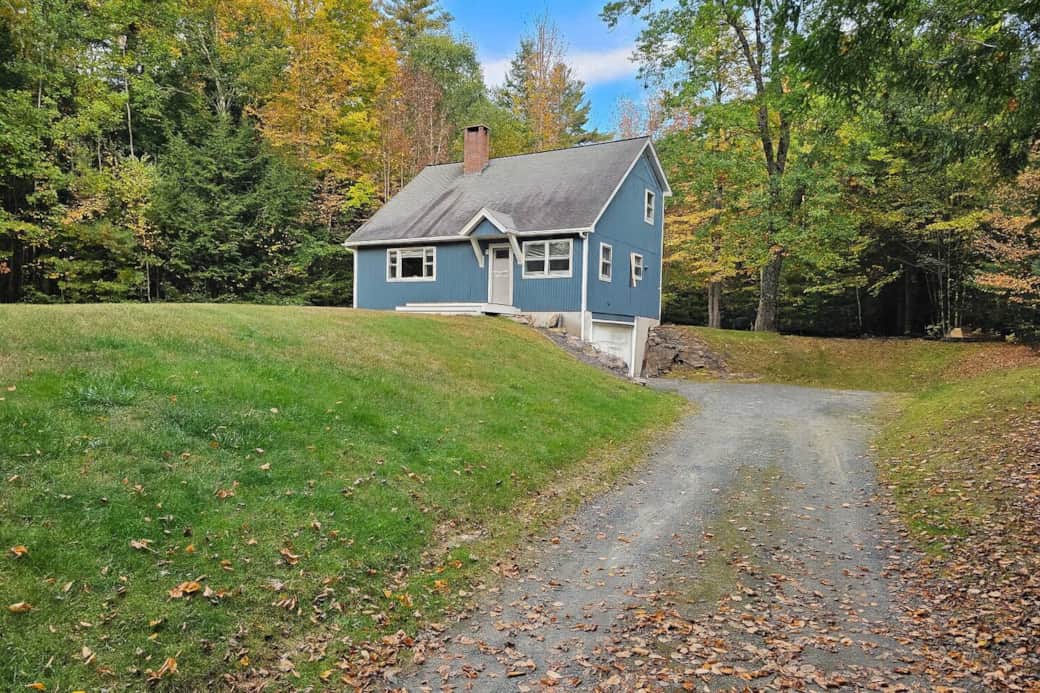 Arrive at your private woodland retreat! This charming blue home welcomes you with a winding driveway, lush lawn, and forest backdrop—your cozy escape in the White Mountains.