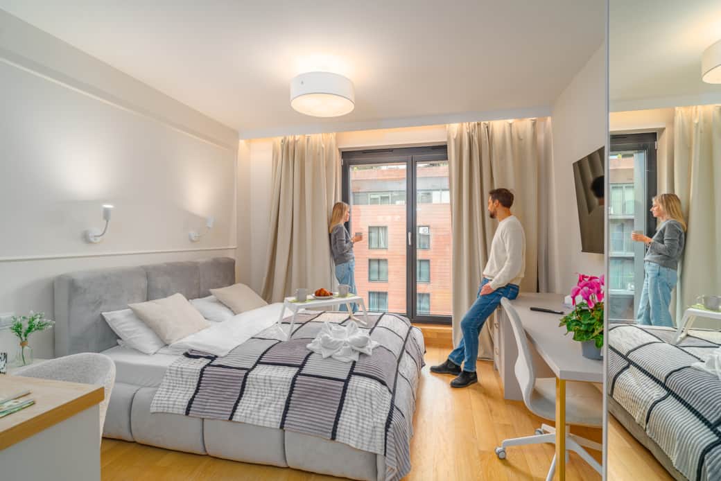 A bright, modern hotel room with a large bed, a desk, a TV, and a balcony.